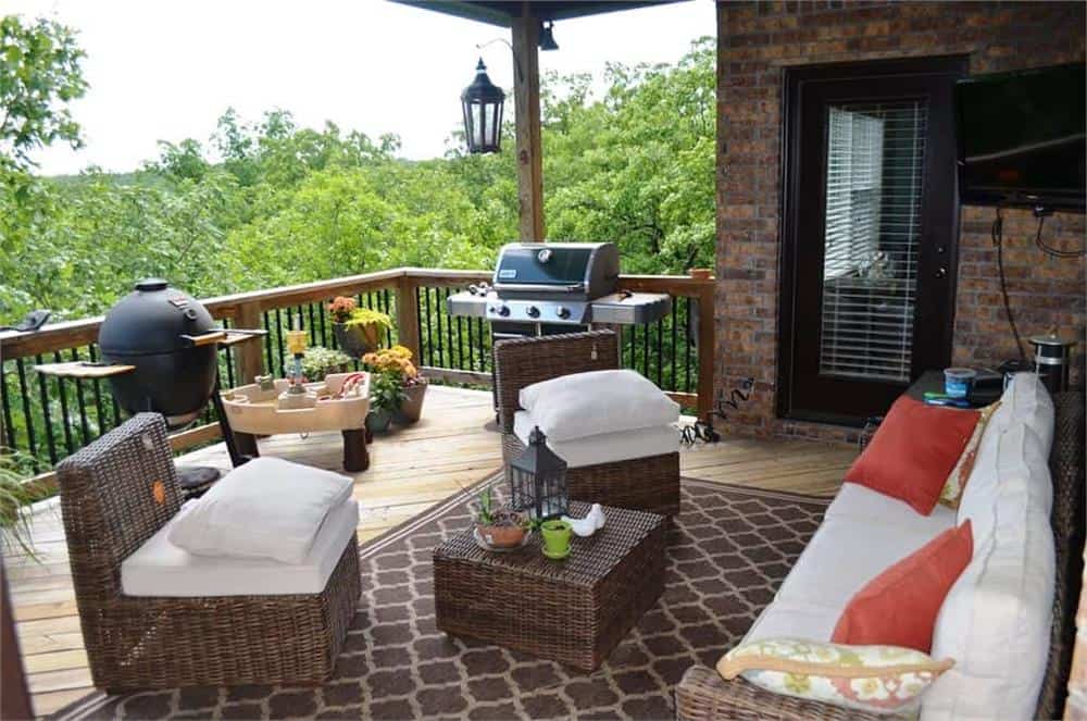 Covered veranda with wicker seats, a matching coffee table, a grill, and wooden railings overlooking the lush greenery.