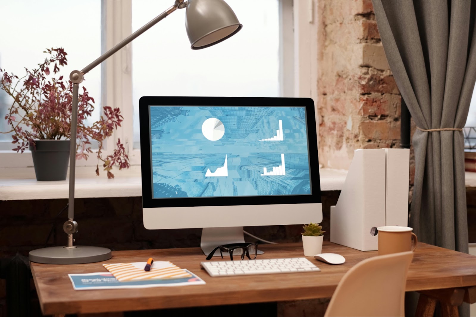 Home office with a computer, keyboard, mouse, plant, cup, and file organizers on a wooden desk, and a lamp on the windowsill.