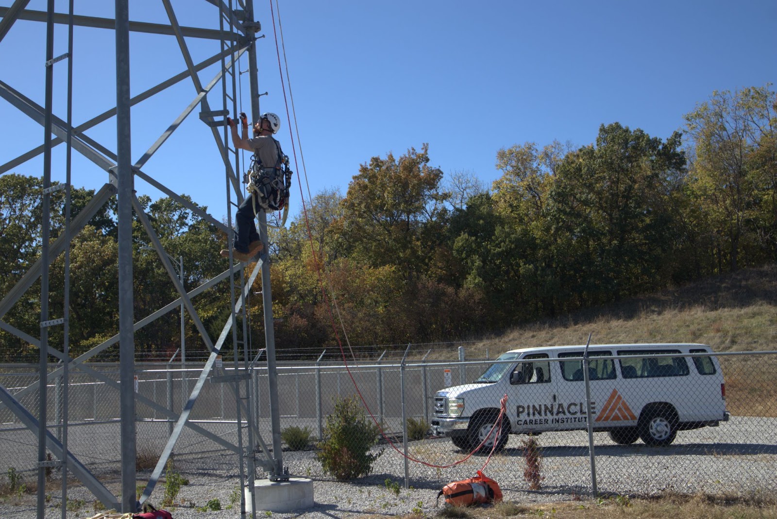 A student in climbing gear is scaling a tower on a sunny day, showcasing hands-on training. A Pinnacle Career Institute-branded van is visible in the background, parked near a fenced area with autumn-colored trees. The image highlights practical education and outdoor technical skills in a professional training setting.