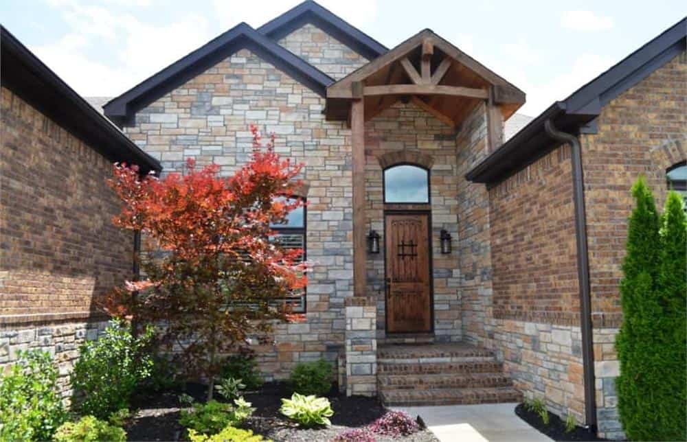 Home entry with a brick stoop, a wooden front door, and a gable roof graced with decorative wood trims.