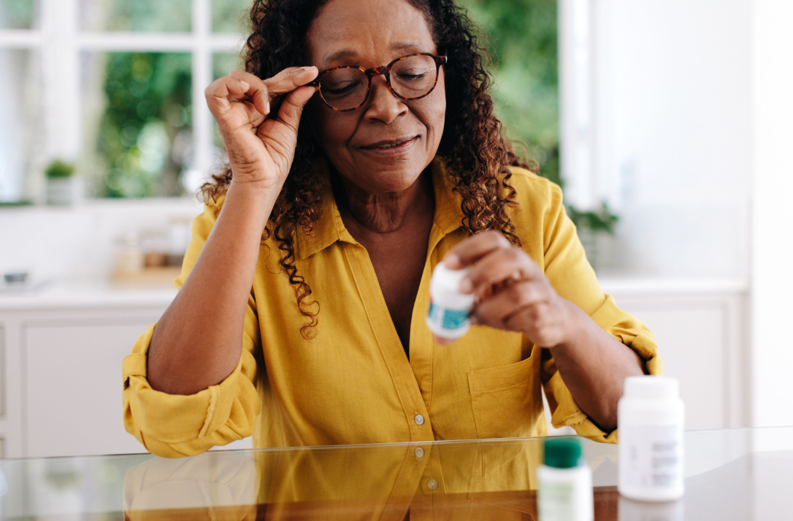 A senior wearing a yellow button-down shirt adjusts their glasses to read a pill bottle label to better understand proper dosage and use.