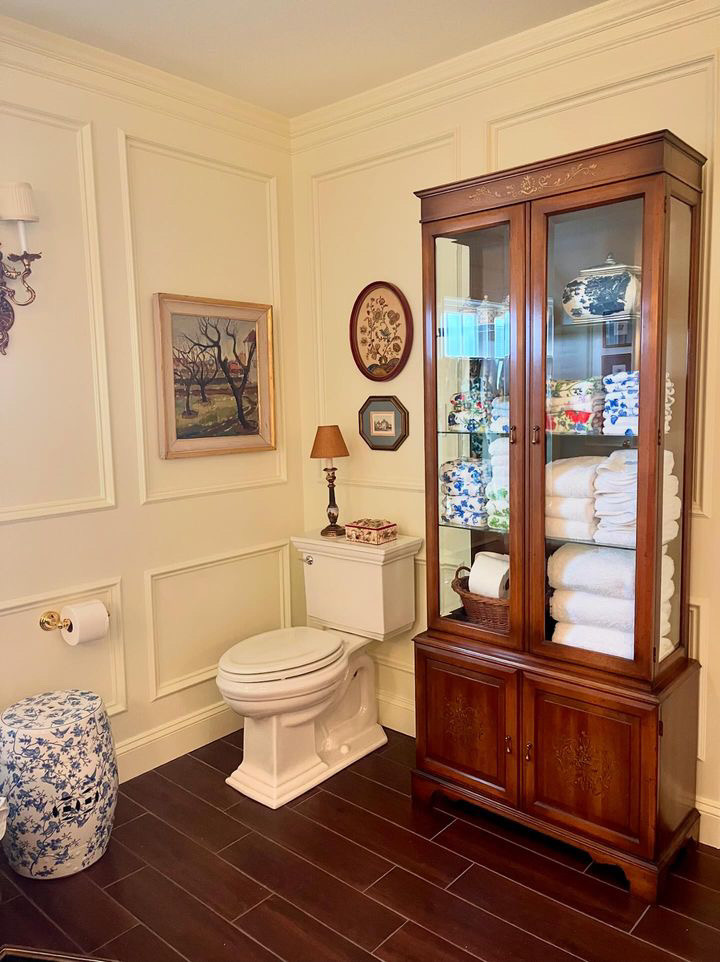 A commode in a bathroom with white paneled walls, featuring a display cabinet filled with linens. 