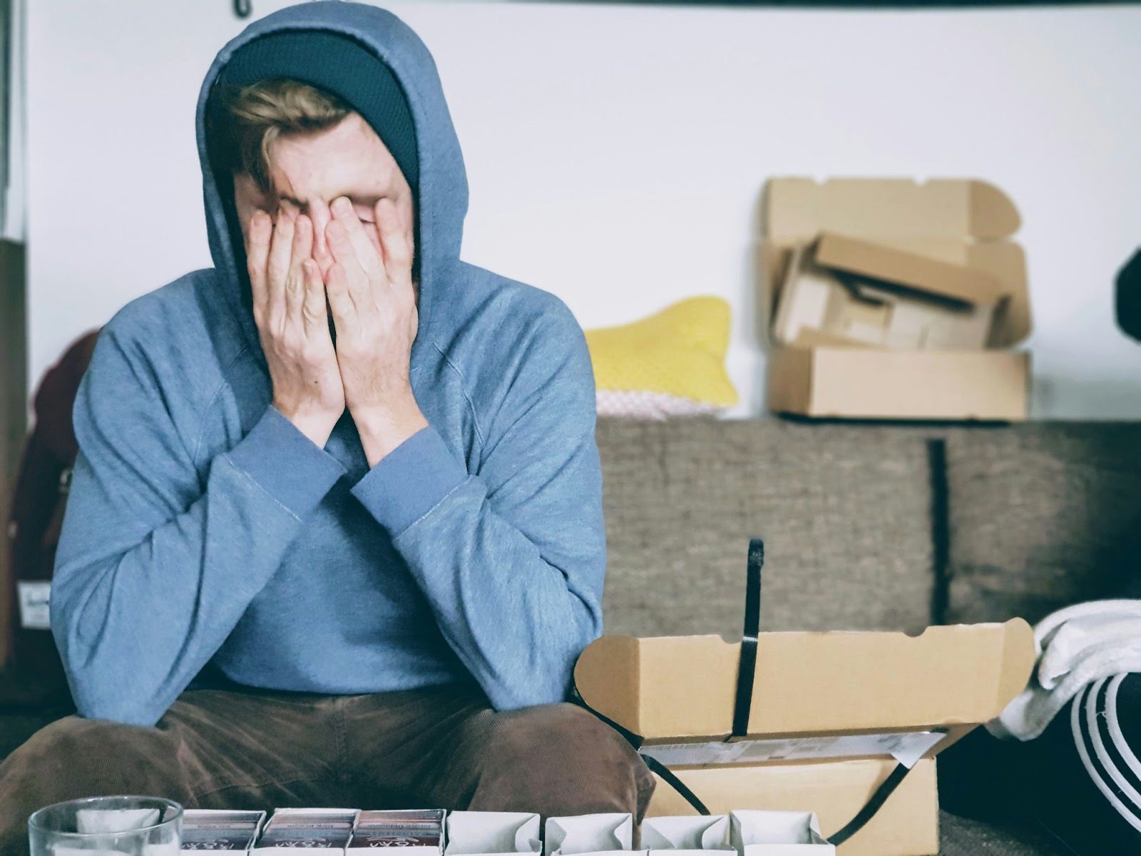 A man sitting on a sofa and covering his face with his hands in the midst of cardboard boxes