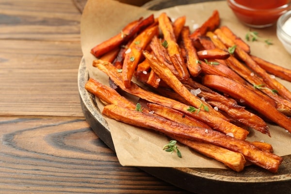 Crispy air-fried sweet potato fries served on parchment paper over a wooden plate, garnished with herbs and sea salt