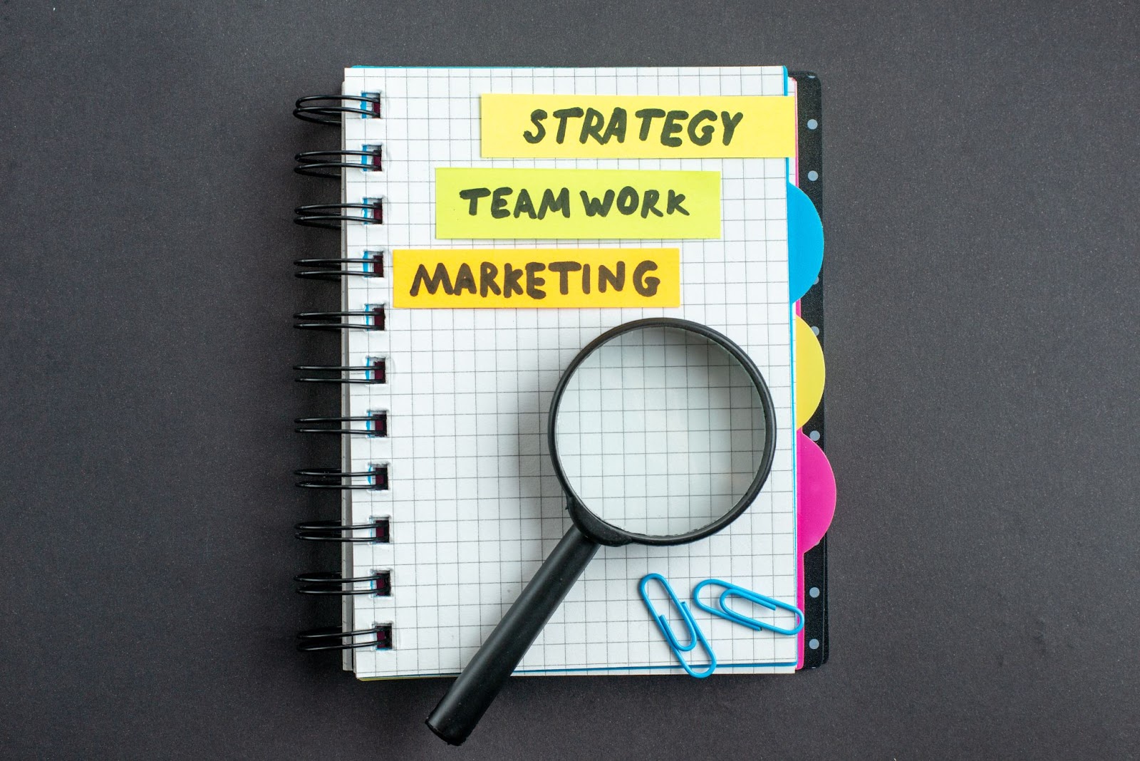 Notebook with the words “strategy,” “team work,” and “marketing” on a notebook next to a magnifying glass and paperclips. 