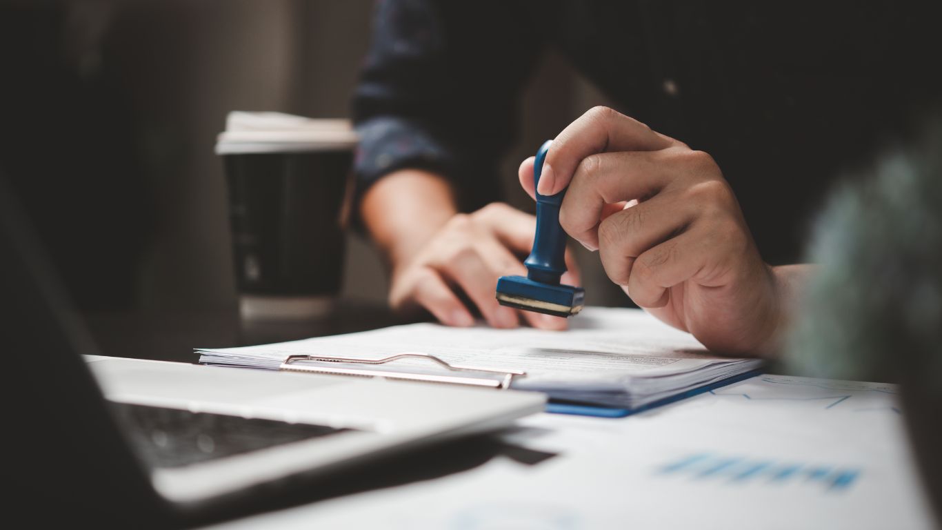  person stamping a document on a desk with a clipboard and paperwork, symbolizing official or legal document processing related to UAE visa amnesty. A coffee cup and laptop are in the background, suggesting an office environment.