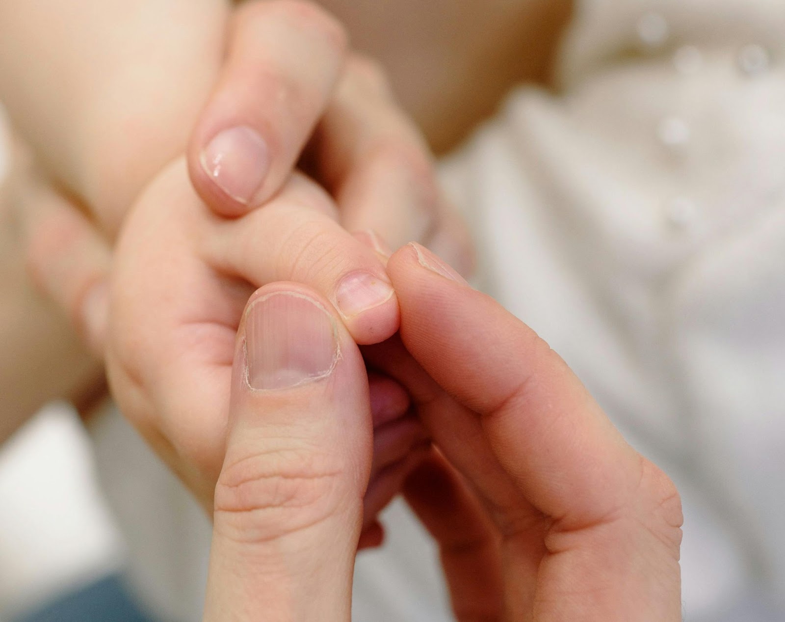 An adult hand uses its thumb and pointer finger to gently pinch a baby thumb finger to show the difference between adult nails and baby nails