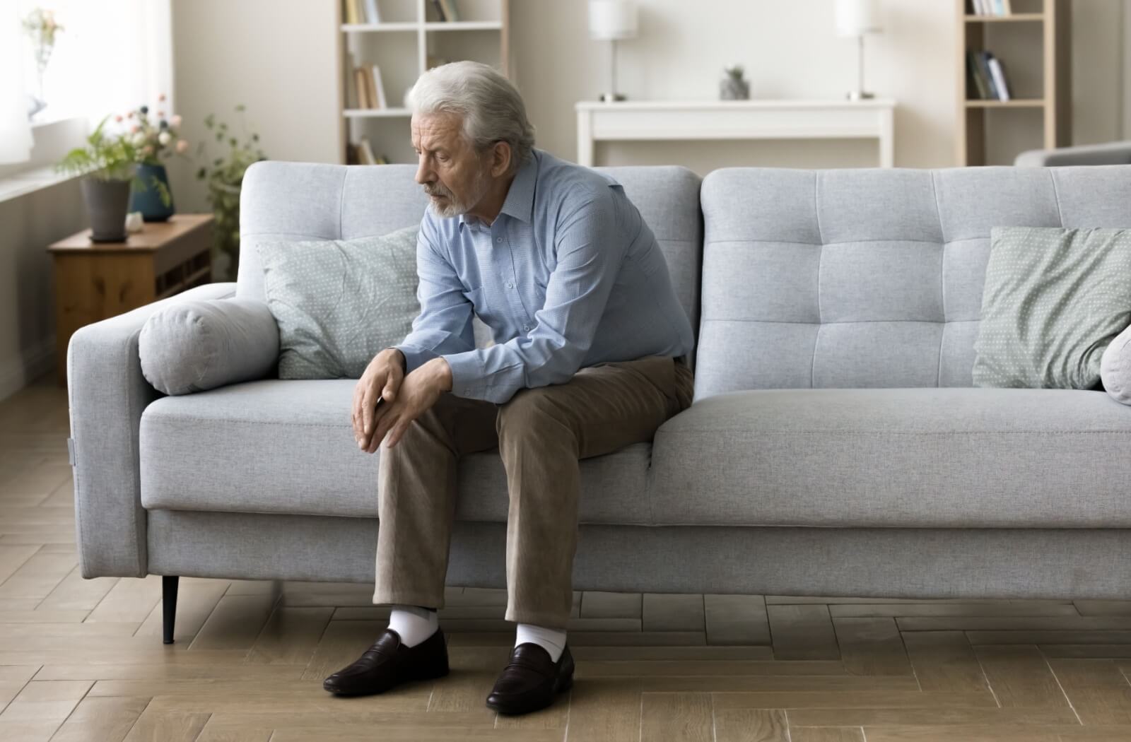 An older adult sits alone on their couch at home, looking pensively at the floor.