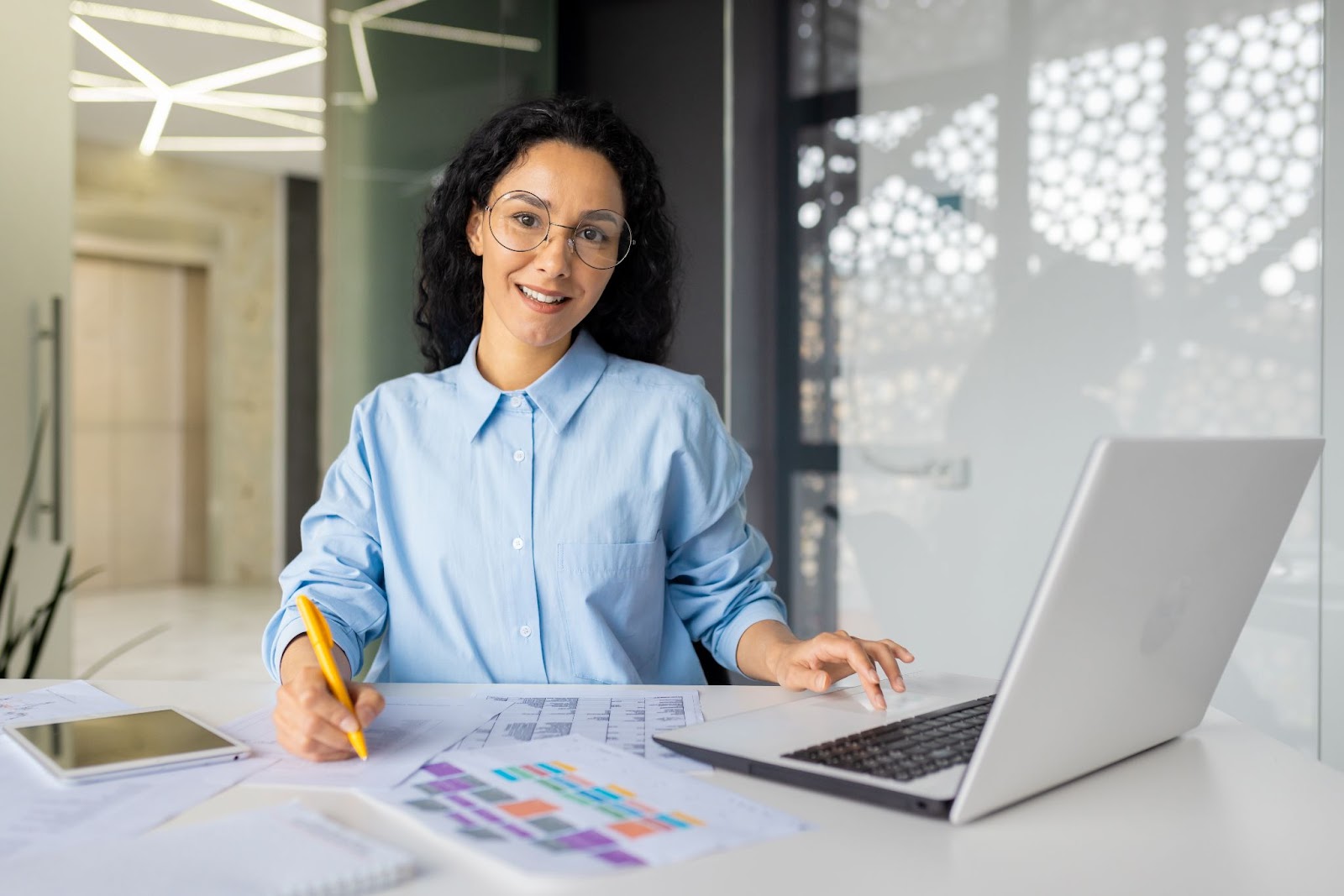 A woman with dark curly hair wears glasses and a blue button-down long-sleeve shirt in a business office. She is working on documents and a laptop and smiling at the camera.