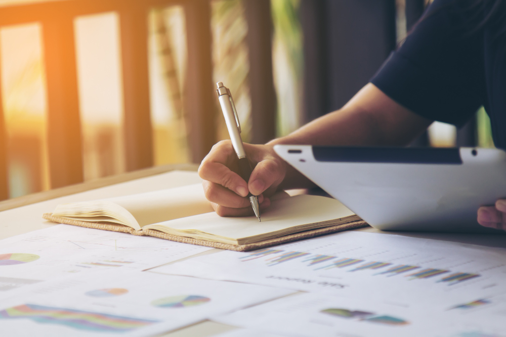 A person is jotting down study tips in a notebook while holding a tablet, surrounded by printed charts and graphs on the table. Sunlight streams through a balcony railing, creating the perfect ambiance for preparing for GATE Architecture 2024.