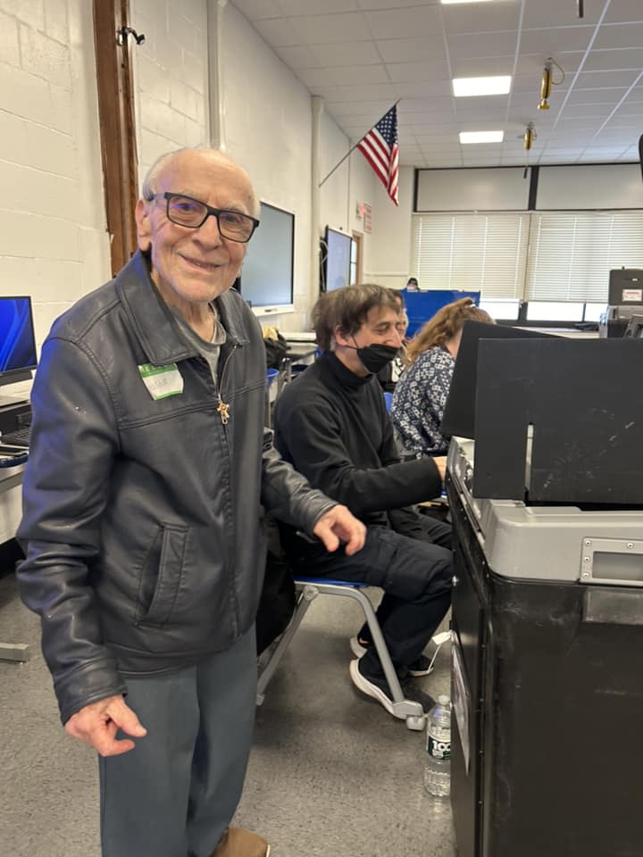 A senior citizen standing and smiling at a camera with people on computers in the background