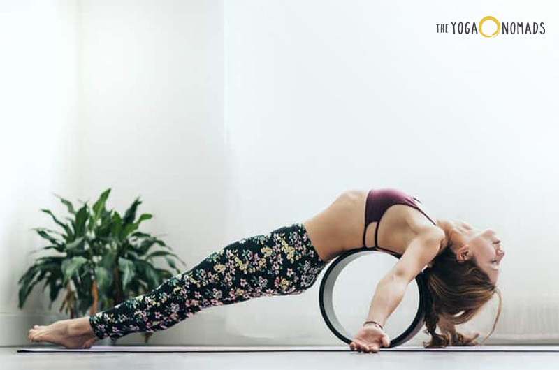 An individual in a fish position using a yoga wheel, which is placed under their back. The background is a white and gray..