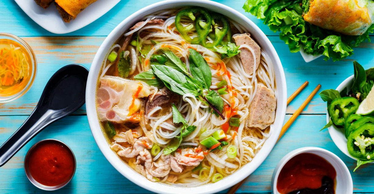 (Eine farbenfrohe Schüssel Pho voller Kräuter, Fleisch, Nudeln und Gewürze – serviert mit frischen Beilagen und kräftigen Saucen für die perfekte, individuell gestaltbare Mahlzeit.)