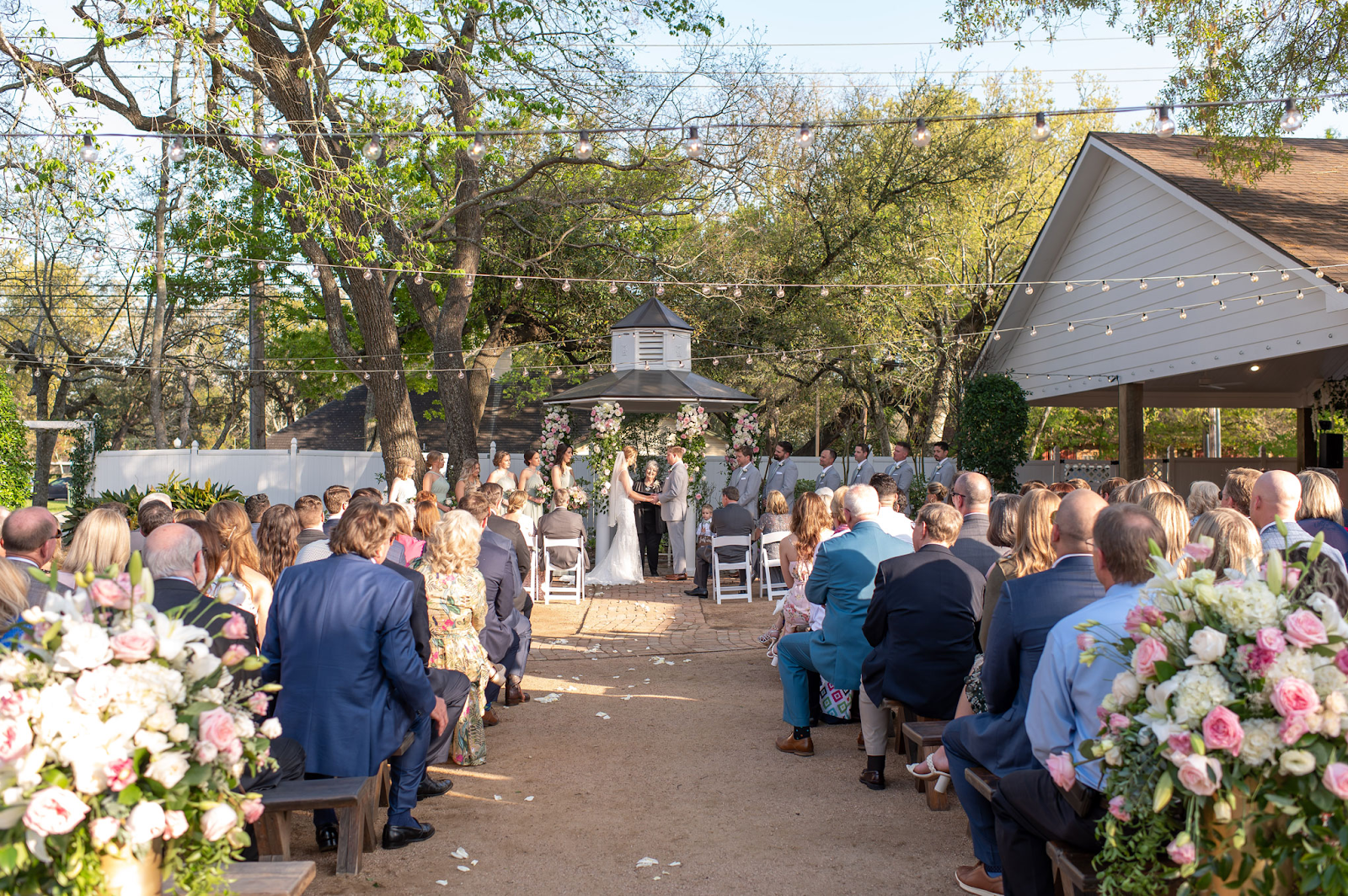 Bride and groom exchange vows under a beautifully adorned gazebo at Butler's Courtyard near Houston Texas, surrounded by lush gardens and stunning floral arrangements created by Lush Flowers. Guests smile and enjoy the intimate outdoor ceremony, enveloped in the romantic and picturesque setting.