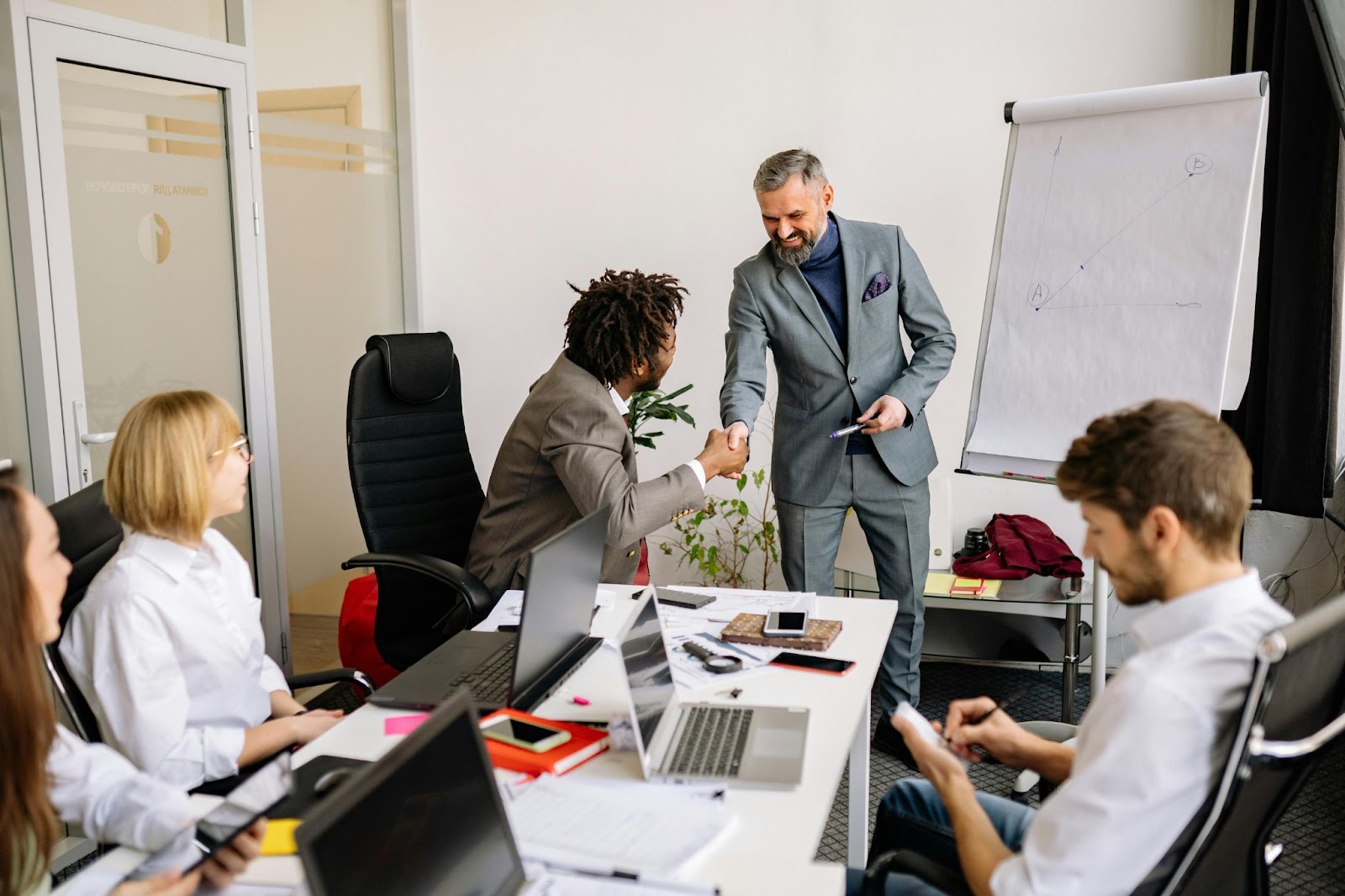 A business meeting where a man in a suit shakes hands with a seated colleague while others work at the table.