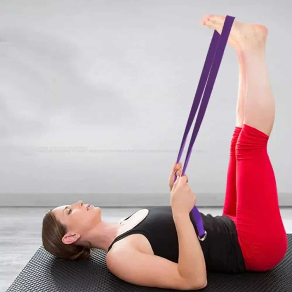 Foto de uma mulher de calça vermelha e camisa regata preta fazendo yoga com uma faixa de yoga roxa deitada de costas em um estúdio com as pernas para cima usando a faixa nos pés e segurando com as mãos.