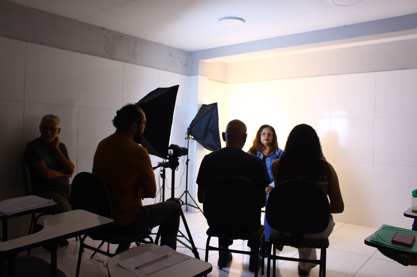 Formação em entrevistas de história de vida do "Nosso Chão, Nossa História", projeto de reparação do desastre socioambiental em Maceió. Uma mulher está sentada, com um refletor no rosto. A sua frente, há pessoas sentadas.

