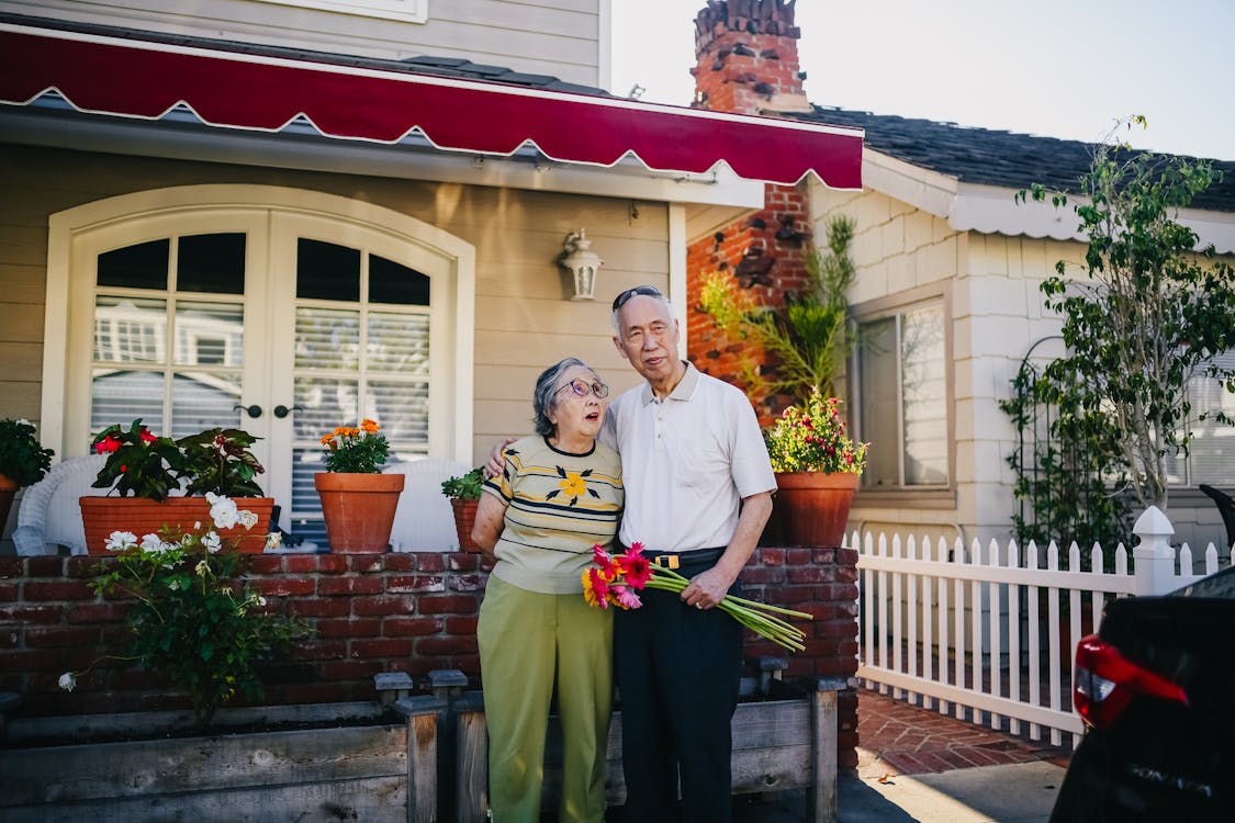 Free Elderly Asian couple embracing outside their cozy home with flowers, representing love and togetherness. Stock Photo