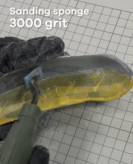 A gloved hand using a 3000-grit sanding sponge and a pen sander to refine a 3D-printed transparent resin model, achieving a smooth, near-glass finish.