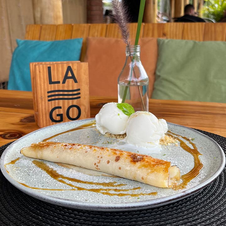 Prato de sobremesa com sorvete de creme decorado com hortelã e calda, servido com uma panqueca doce ao lado, em ambiente de restaurante aconchegante.