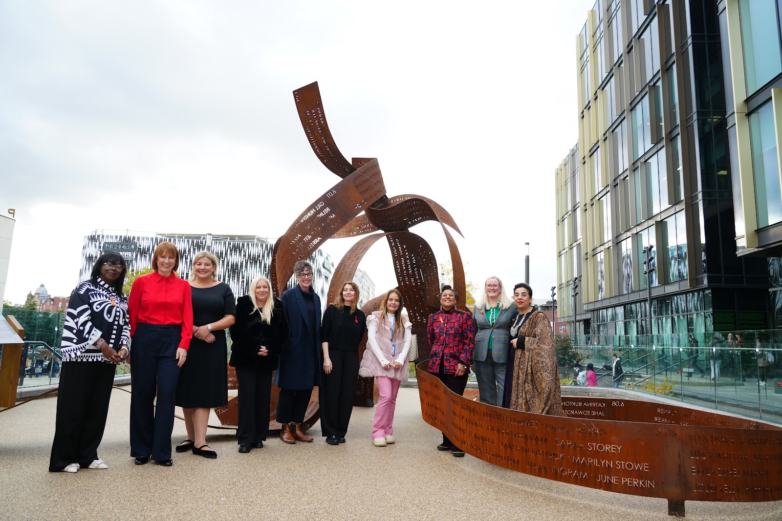 ten women standing in a line in front of a large sculpture of metal ribbons