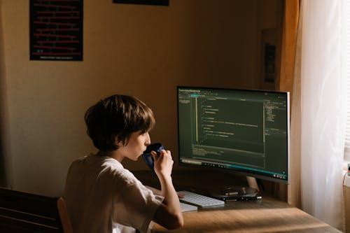 Free A young person coding at a desk with a computer and drinking from a mug. Stock Photo