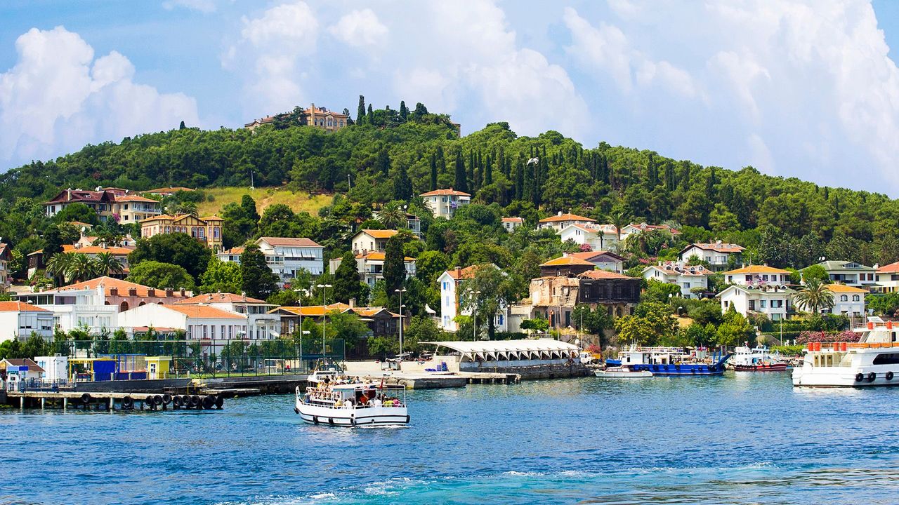 Scenic view of Adalar (Princes' Islands) near Istanbul, Turkey, with colorful houses, lush green hills, and boats sailing on the clear blue waters of the Marmara Sea.