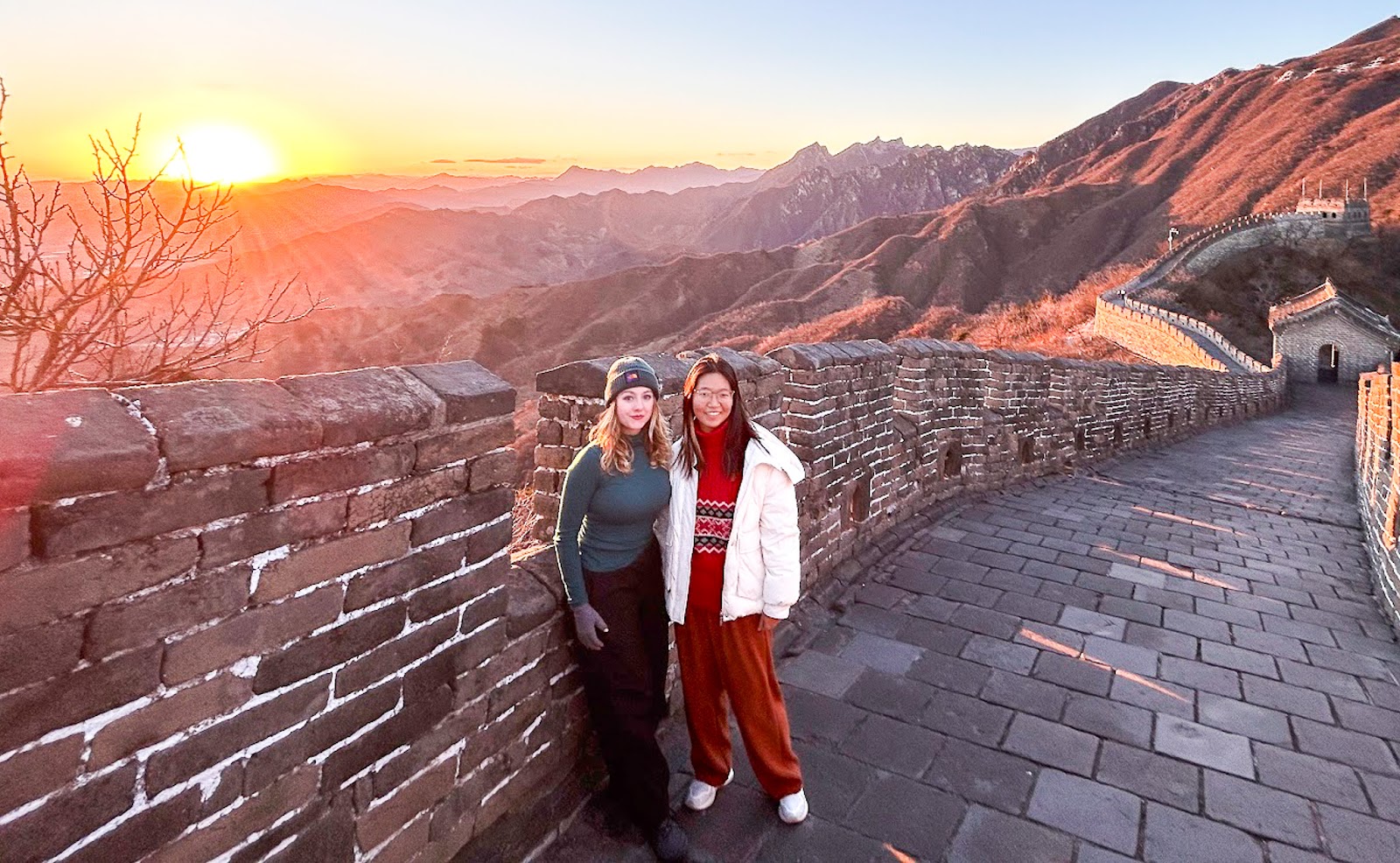 two girls stand next to each other on the Great Wall while the sun sets behind them