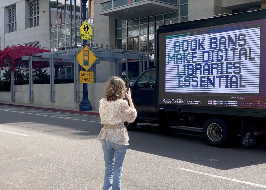 A person stands in the street to take a photo of a truck with a large screen on the side. It reads "book bans make digital libraries essential."