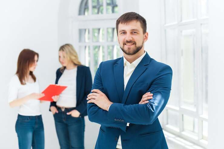 A man smiling on the foreground while two females stand in the background and discuss something