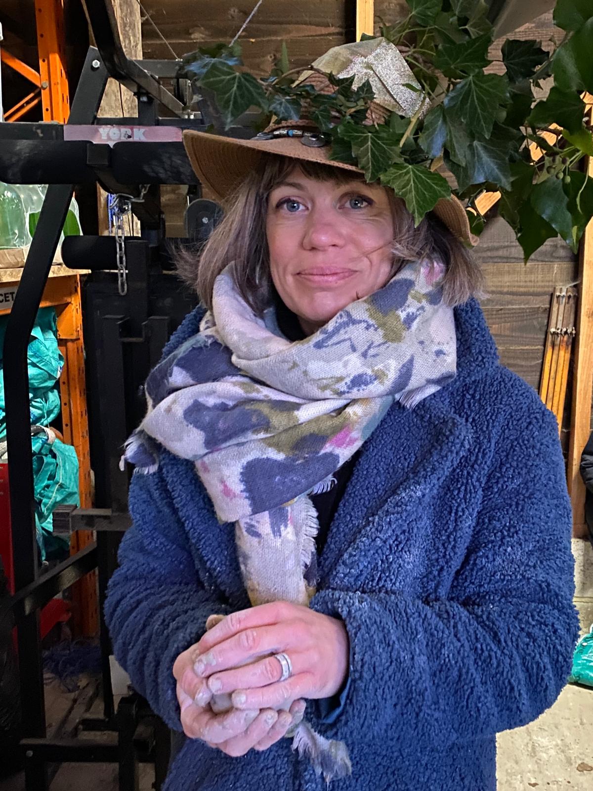 A white woman with shoulder length blonde hair and an ivy adorned hat wears a large floral scarf and blue fleece jacket. You can see her hands are covered in clay chalk as she holds a lump of clay.