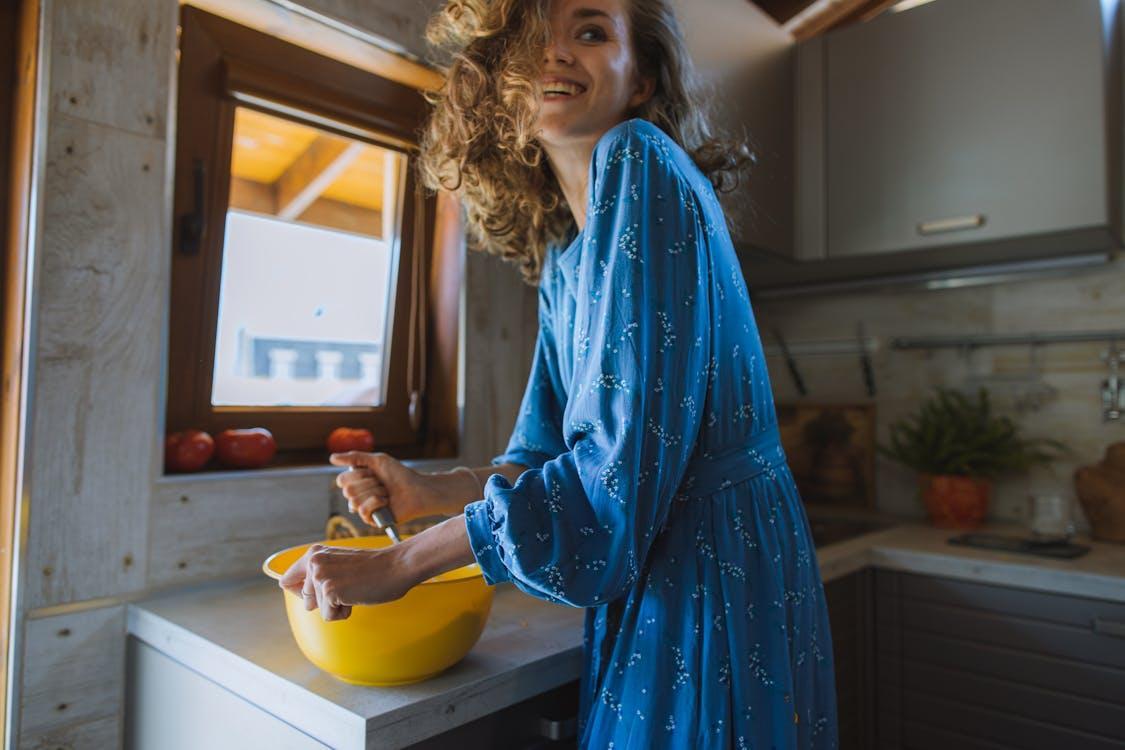 Free A smiling woman in a blue dress prepares food beside a window, enjoying her indoor cooking experience. Stock Photo