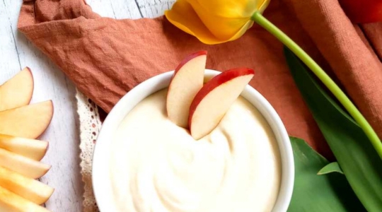 Un bol de trempette au yaourt crémeux avec des tranches de pomme fraîches sur le côté, placé à côté d'une tulipe jaune sur une surface en bois.