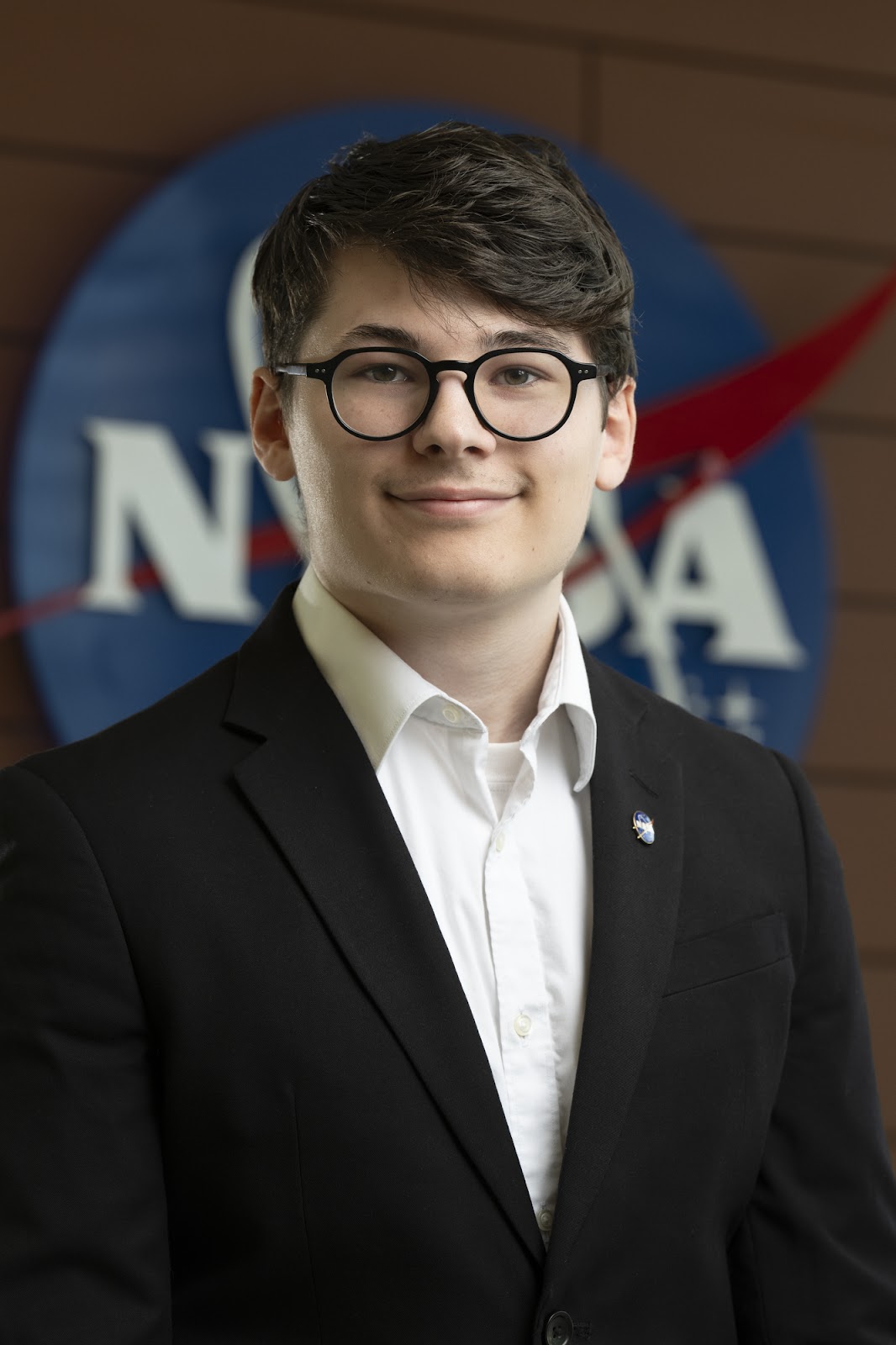 Logan Buttler, college student wearing glasses and a suit in front of NASA insignia.