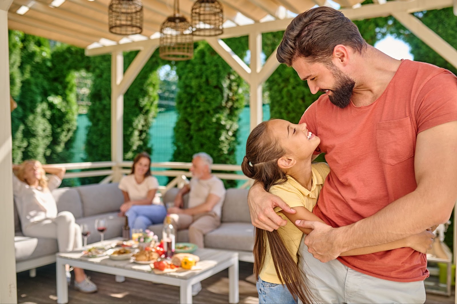 Man hugging his daughter under a vinyl patio cover with three people sitting on a couch in the background.