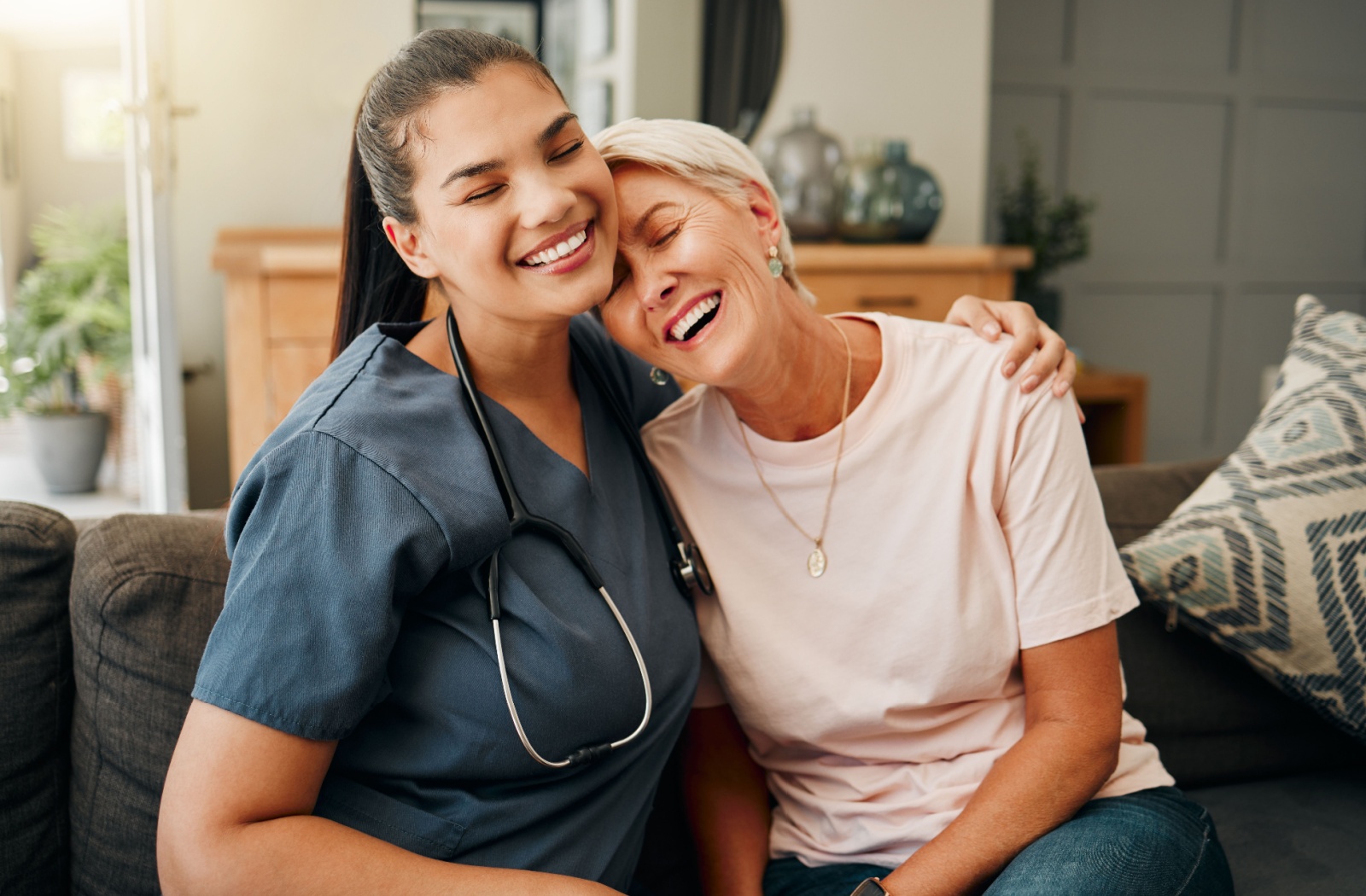 A caregiver and a senior woman smiling and hugging on the couch in respite care.