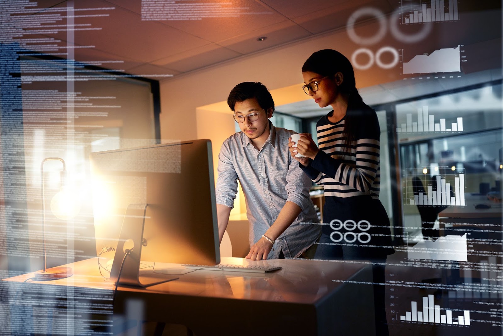 Two colleagues collaborate on a user interface in a dimly lit office at night, with reflections from multiple screens in the foreground.