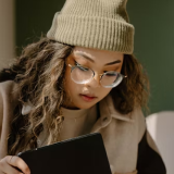 A person wearing a beanie and glasses reads a book or tablet indoors.