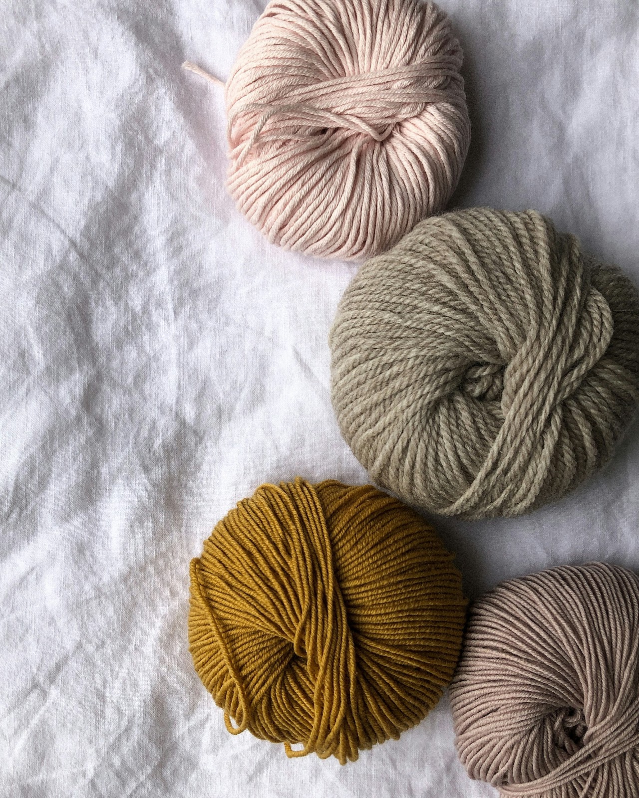 Four balls of merino wool  in soft earthy tones—pink, beige, mustard, and taupe—on a white fabric background..