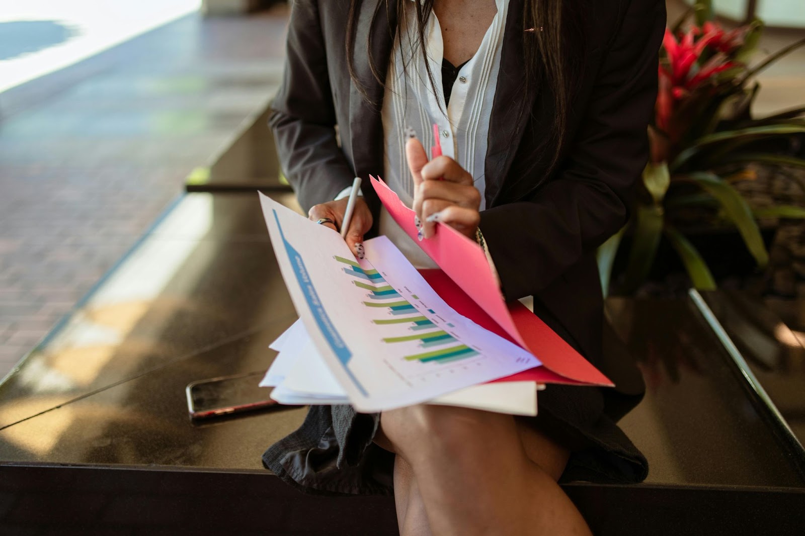 A person holding documents with graphs and charts on them, seated on a bench with a phone next to them.