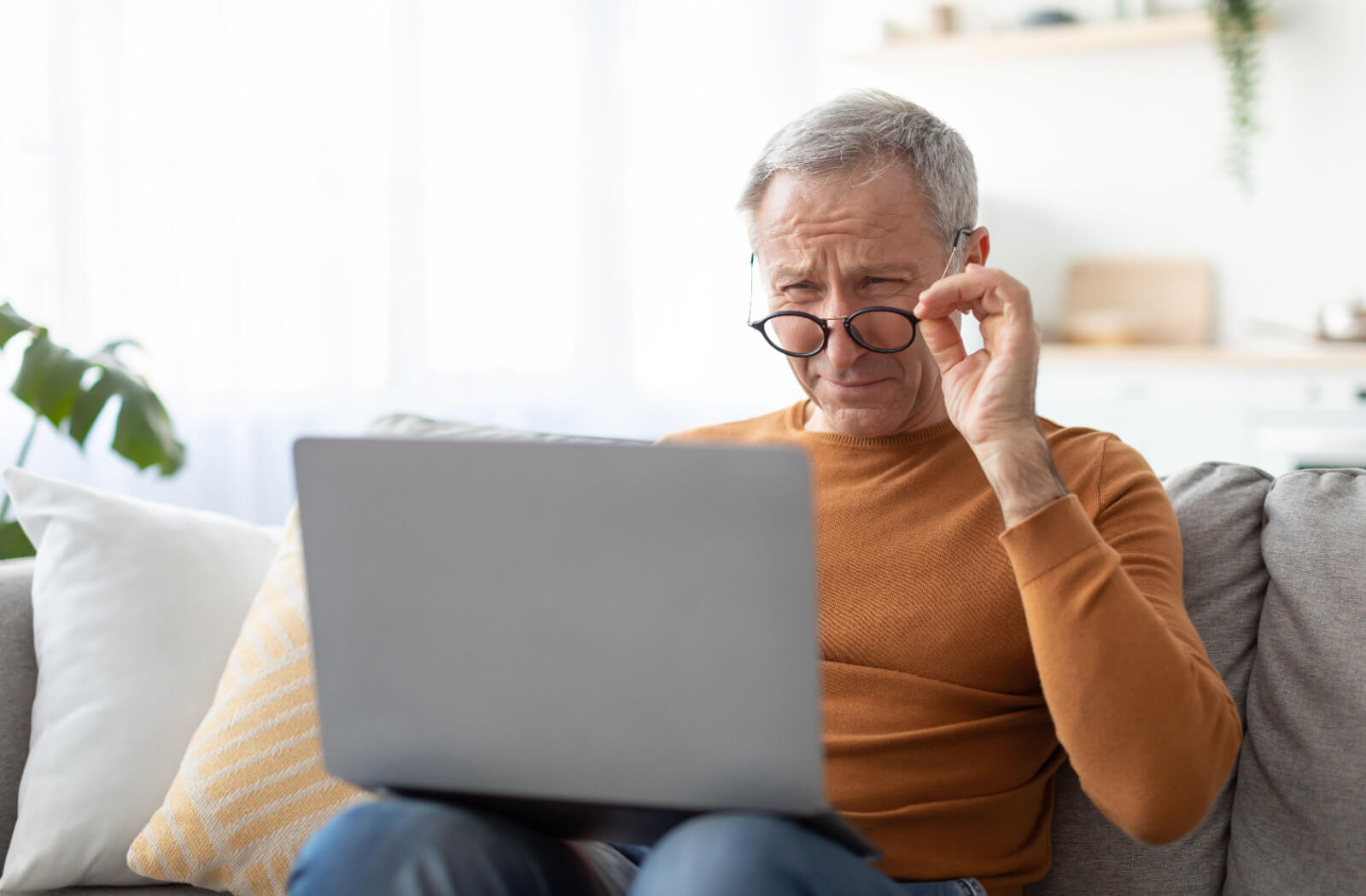 A senior man sitting on the couch pulling down his glasses to focus on his computer due to his vision getting worse.