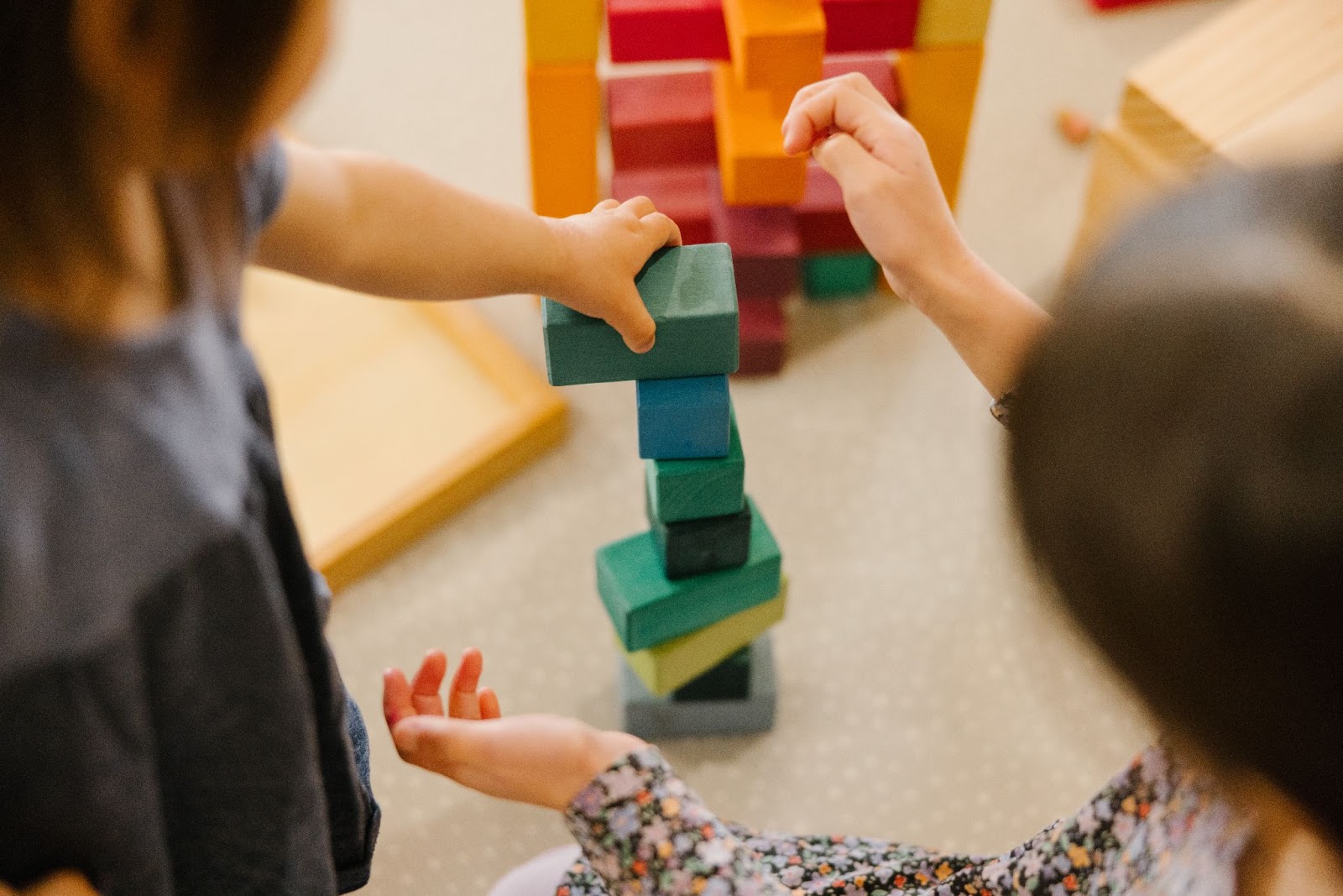 Two children carefully and meticulously stack the vibrant Grimm’s Large Stepped Pyramid blocks, selecting similar color tones to create their imaginative structures.
