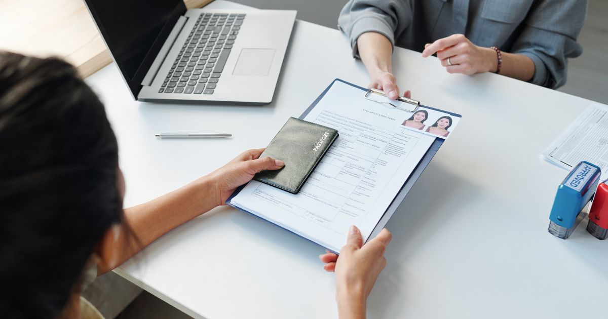 A woman handing over a passport and a completed application form on a clipboard across a table, with another person receiving the documents, likely in an official setting such as a visa application or interview.