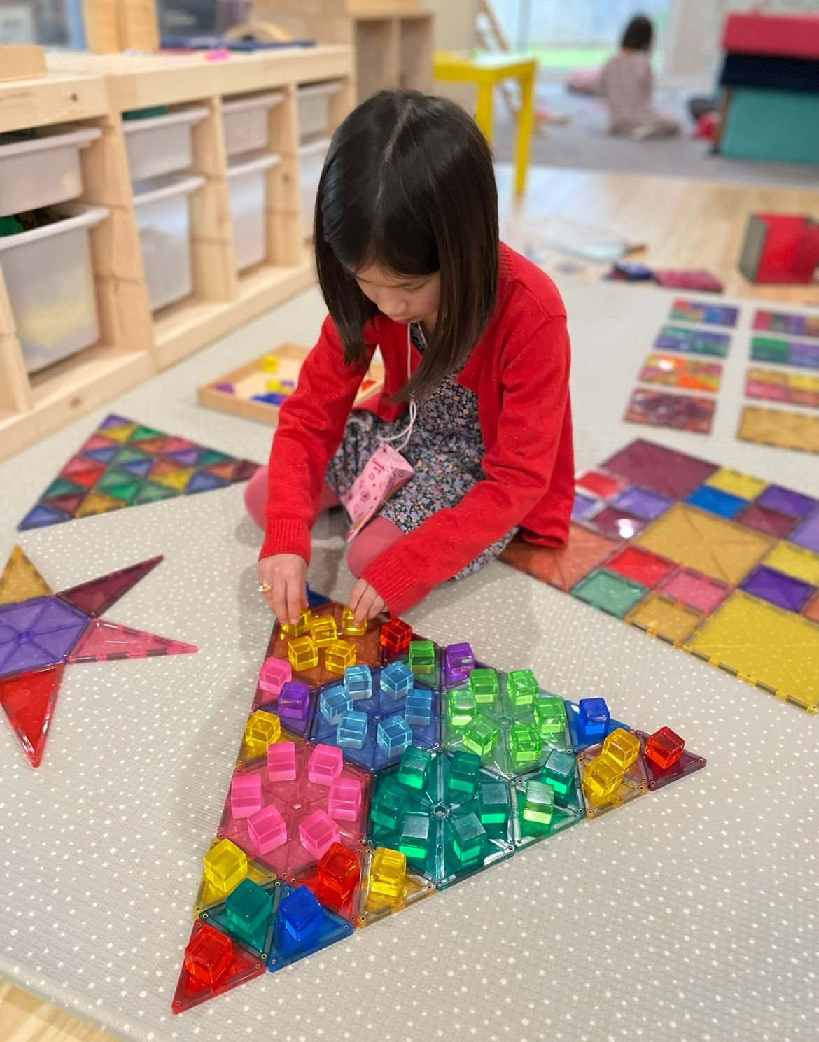 A preschooler is arranging colourful acrylic blocks on a large triangle made from magnetic tiles. The child is engaged in creative play, carefully placing the blocks to match the vibrant colours of the tiles, showcasing their imagination and focus.
