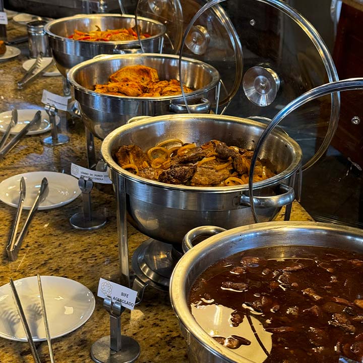 Buffet de comidas variadas com pratos quentes em panelas de aço inox, incluindo carne, molho e legume, em um evento ou restaurante, atraente e bem organizado.