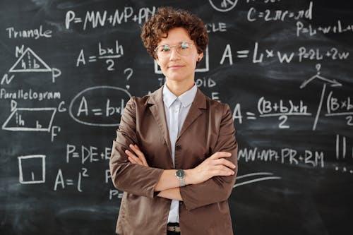 Free Confident female teacher standing with arms crossed in front of a detailed mathematical blackboard. Stock Photo