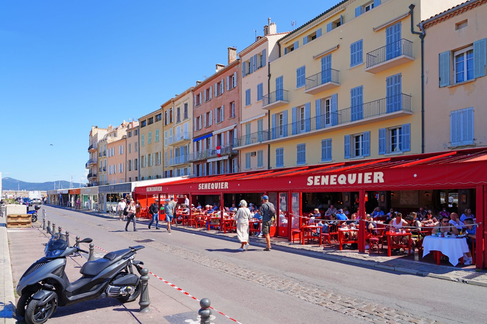 Café Sénéquier in Saint-Tropez with its signature red awning and outdoor seating filled with guests along a pastel-colored building-lined waterfront street under a clear blue sky.
