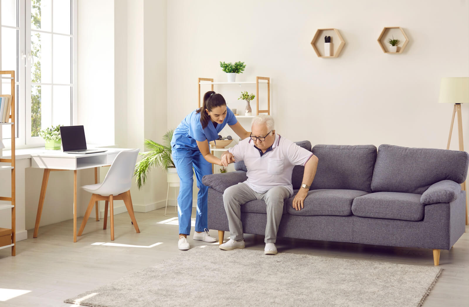 A caregiver bending over and helping an older adult stand from his couch in his new assisted living community.