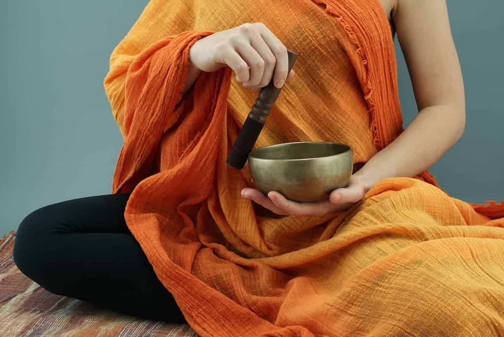 A person seated in a meditative pose, wearing an orange robe, with one hand holding a wooden mallet and the other hand holding a Tibetan singing bowl. The setting suggests mindfulness or meditation practice.