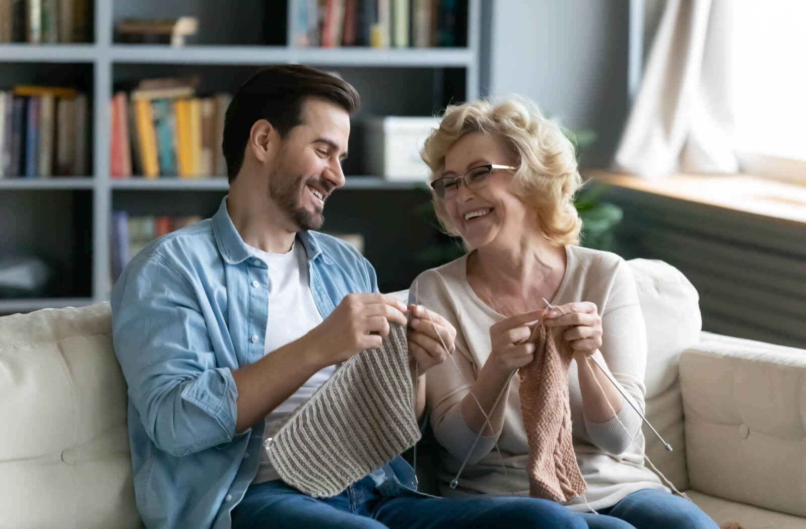 A senior woman laughing while teaching her adult son how to knit during arts and crafts.